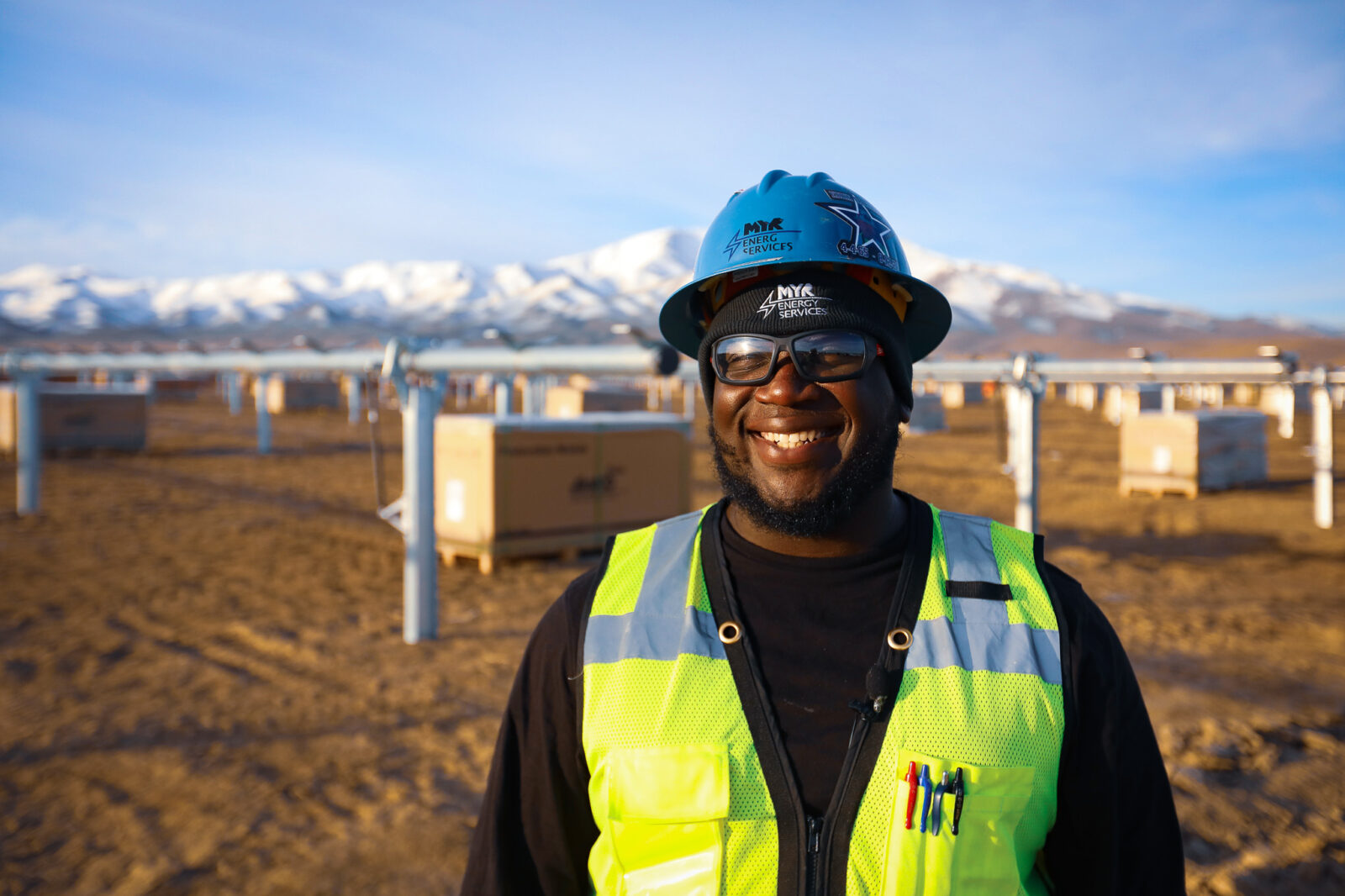 MYRE Energy employee with hard hat and safety glasses smiles for the camera with snowcapped mountains in the distance