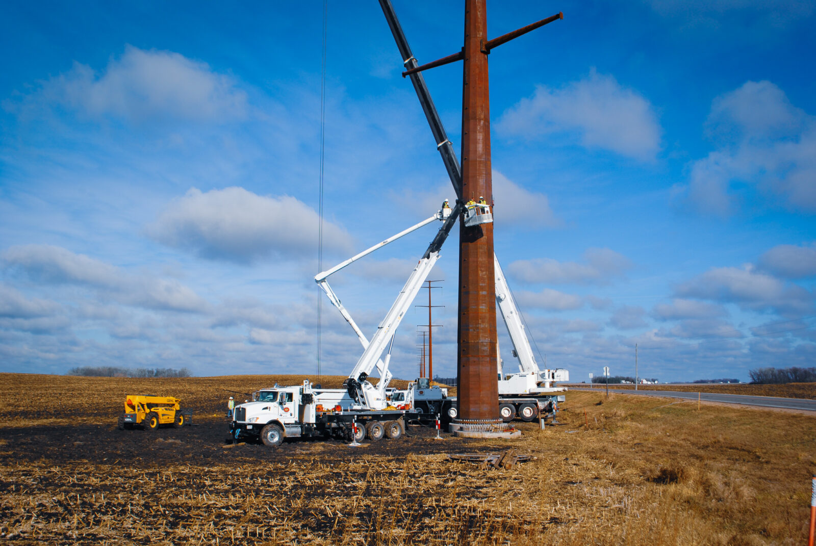 Two white bucket trucks elevate L.E. Myers employees for transmission line work in brown field