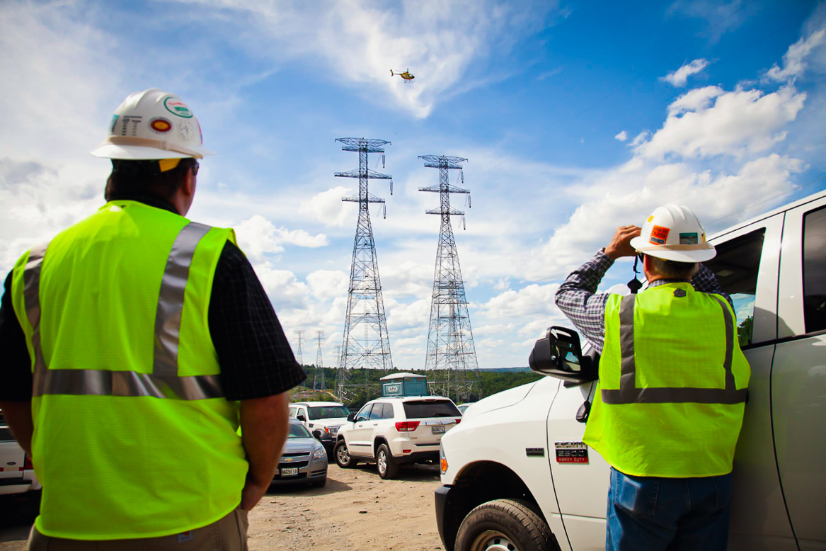 Two MYR Group employees watch helicopter flying over transmission lines through binoculars