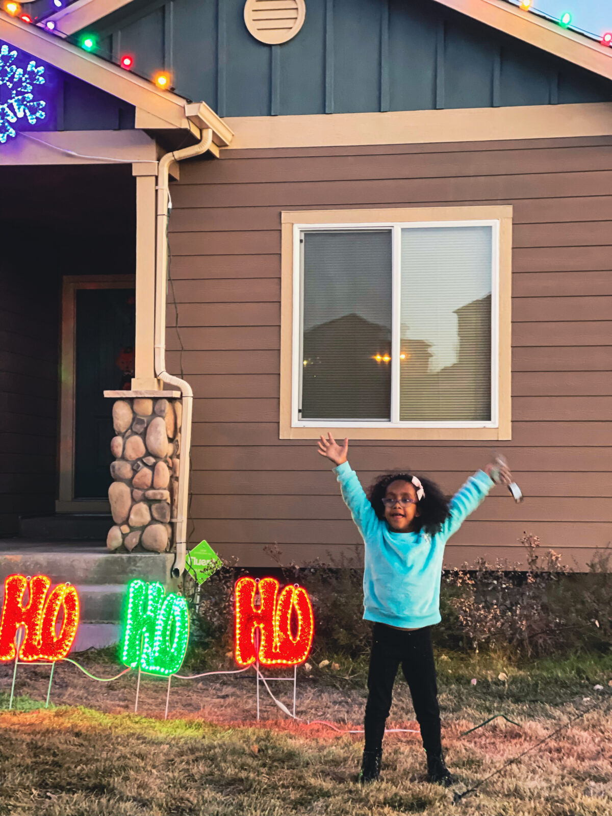 Little girl stands out front of her house, arms raised next to Christmas lights.