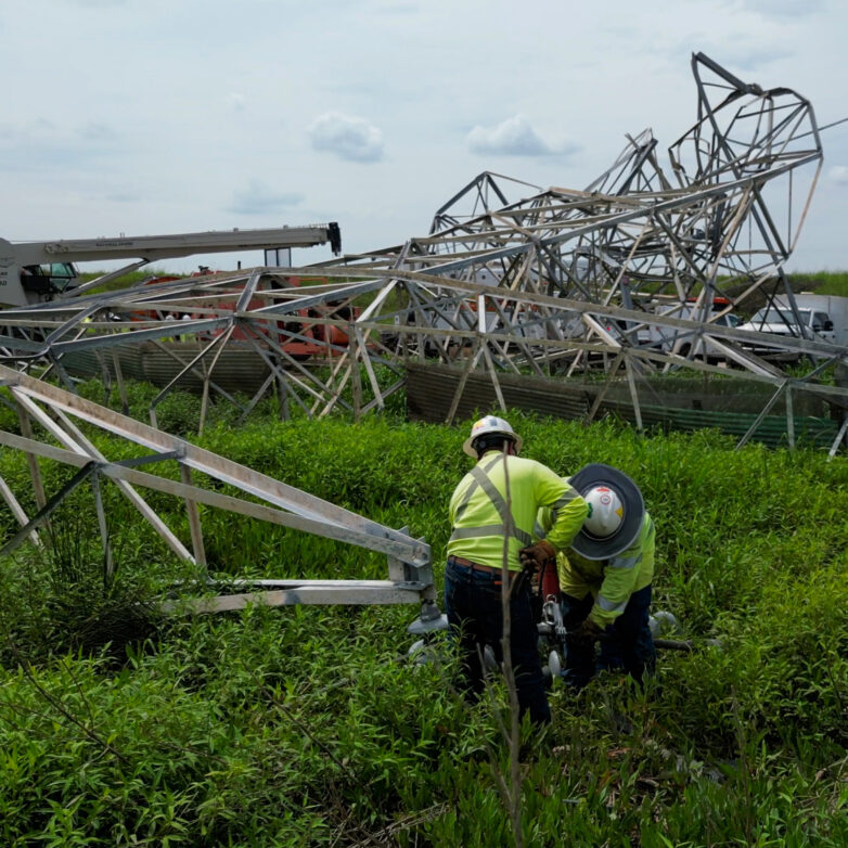 A storm damaged transmission tower lies on the ground. Two construction workers examine a portion of the steel.